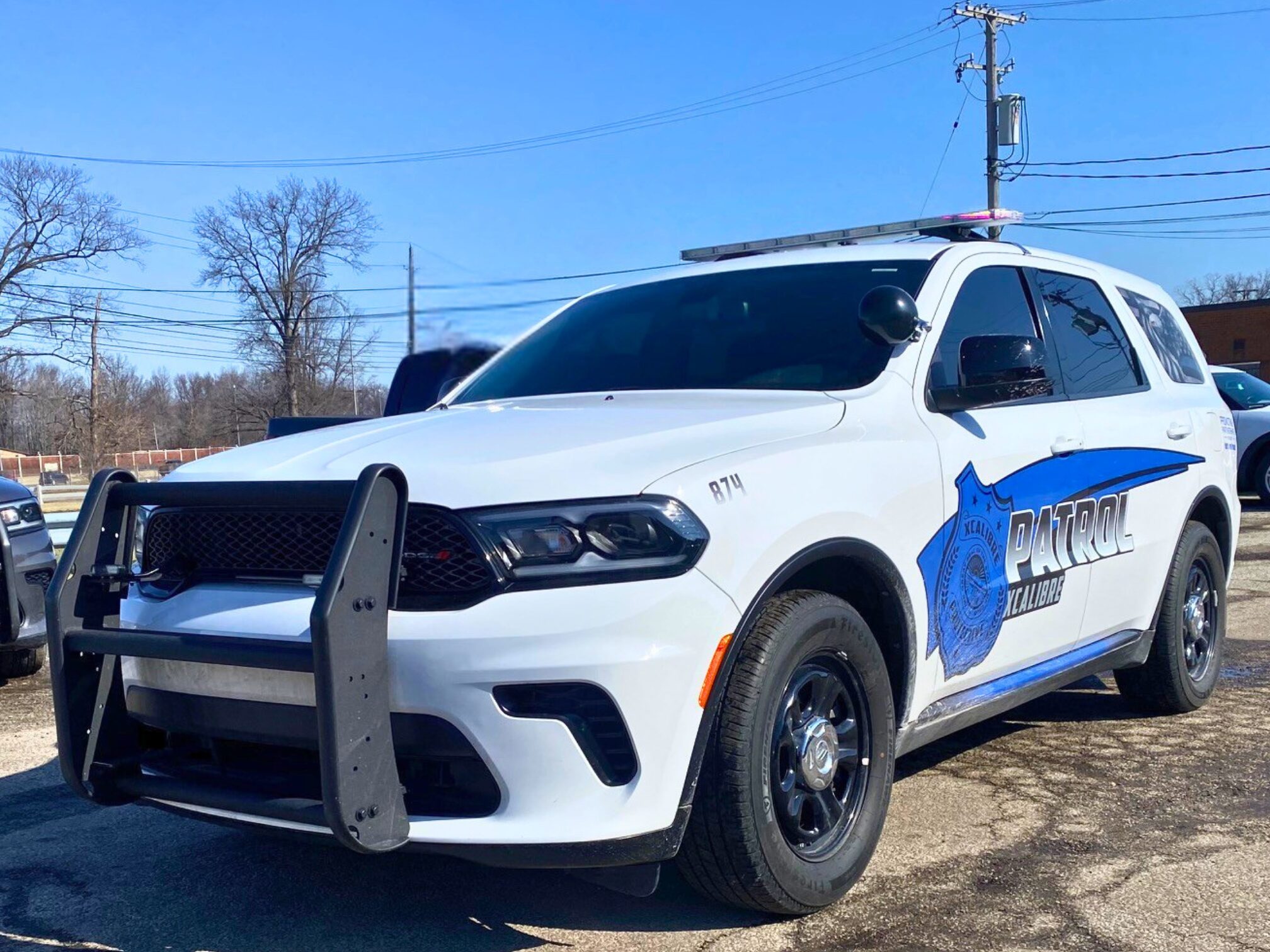 A white Xcalibre Protective Services patrol SUV with a black grille and emergency lights parked in a lot under a clear blue sky.
